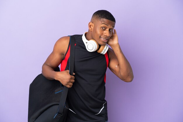 Young sport latin man with sport bag isolated  on purple listening to something by putting hand on the ear