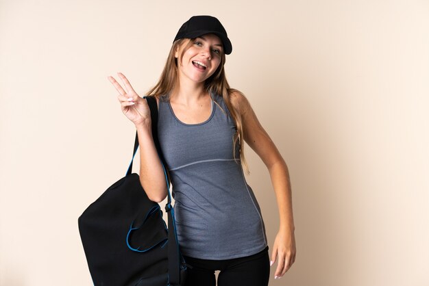 Young sport Lithuanian woman holding a sport bag isolated on beige wall smiling and showing victory sign