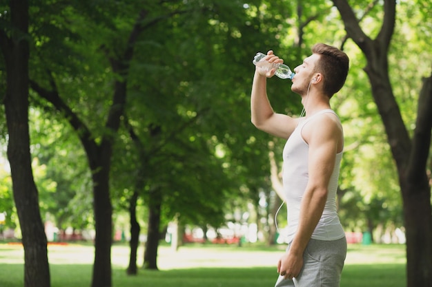 トレーニング後に水を飲む若いスポーツ男。ペットボトルを保持し、公園でのトレーニング後に休息をとって、健康的なライフスタイルの概念のイヤホンのフィットネス男