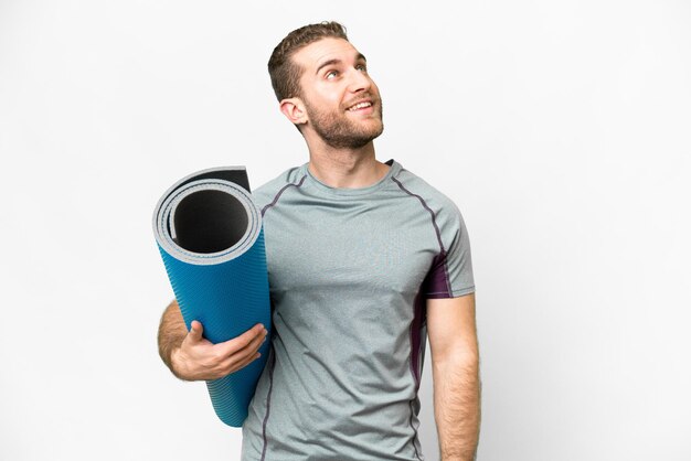 Young sport man going to yoga classes while holding a mat over isolated white background looking up while smiling