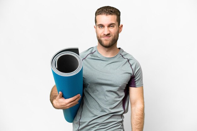 Young sport man going to yoga classes while holding a mat over isolated white background with sad expression