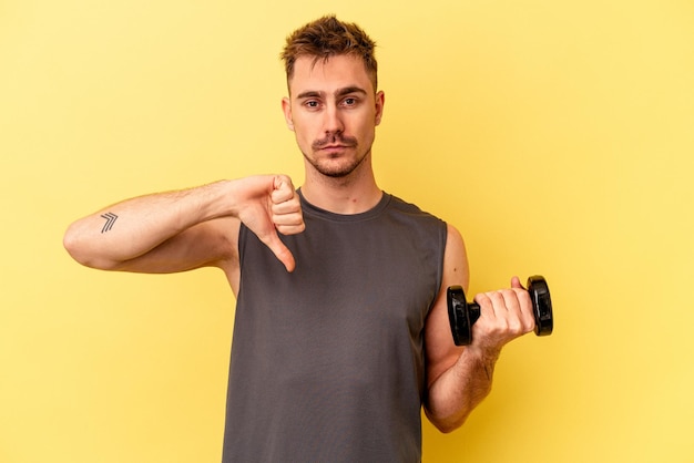 Young sport man holding a dumbbell isolated on yellow background showing a dislike gesture, thumbs down. Disagreement concept.