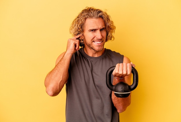 young sport man making weightlifting isolated on yellow background covering ears with hands.