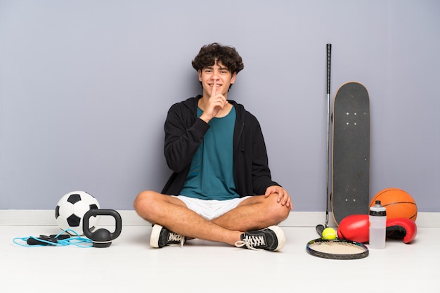 Young sport man sitting on the floor around many sport elements doing silence gesture