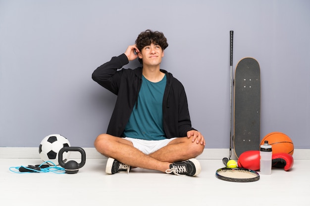 Young sport man sitting on the floor around many sport elements having doubts and with confuse face expression