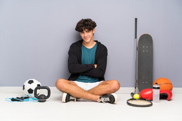 Foto uomo giovane sportivo seduto sul pavimento intorno a molti elementi sportivi ridendo