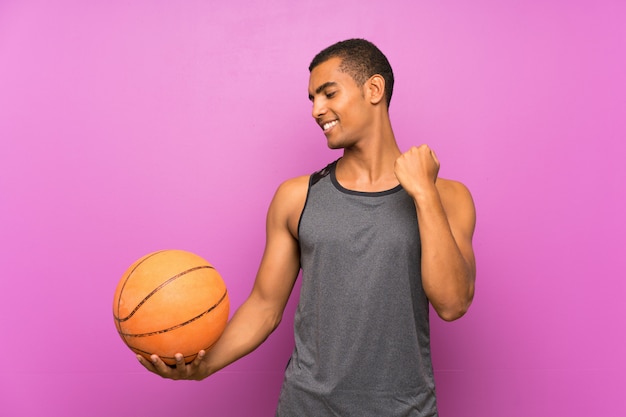 勝利を祝う孤立した紫色の壁の上のバスケットボールのボールを持つ若いスポーツ男