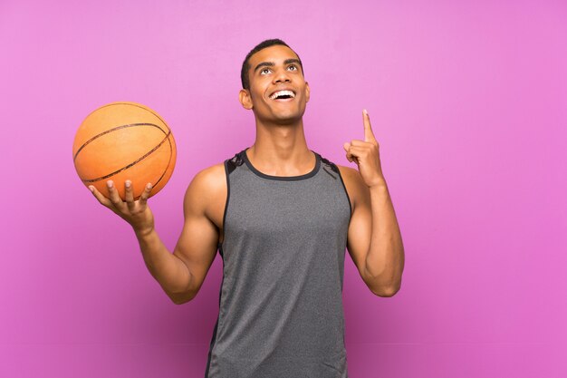 Young sport man with ball of basketball over isolated purple wall pointing up a great idea