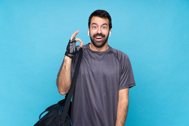 Young sport man with beard over isolated blue  surprised and showing ok sign