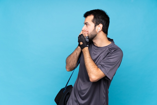 Young sport man with beard over isolated blue wall covering mouth and looking to the side