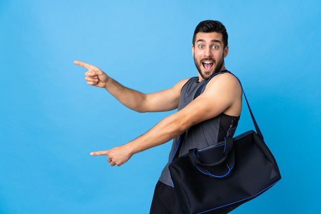 Young sport man with sport bag isolated on blue pointing finger to the side and presenting a product