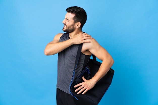 Young sport man with sport bag isolated on blue suffering from pain in shoulder for having made an effort