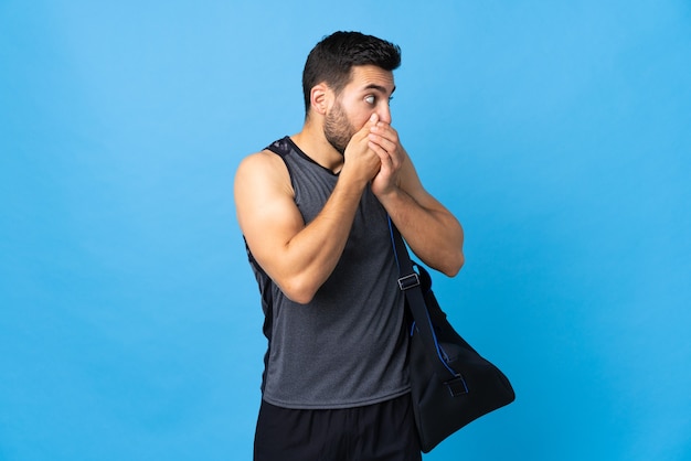 Young sport man with sport bag isolated on blue wall covering mouth and looking to the side