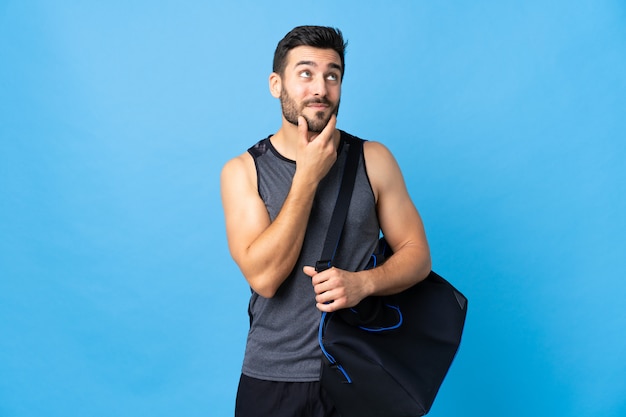 Young sport man with sport bag isolated on blue wall thinking an idea while looking up