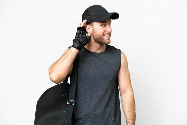 Young sport man with sport bag over isolated white background listening to something by putting hand on the ear