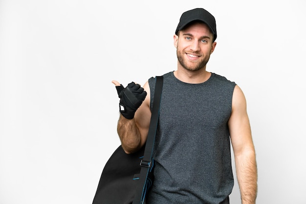 Молодой спортсмен со спортивной сумкой на изолированном белом фоне указывает в сторону, чтобы представить продукт