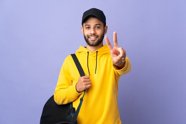 Молодой спортивный марокканский мужчина со спортивной сумкой, изолированной на фиолетовой стене, улыбается и показывает знак победы