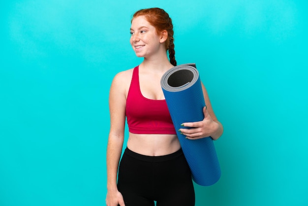 Young sport reddish woman going to yoga classes while holding a mat isolated on blue background looking to the side