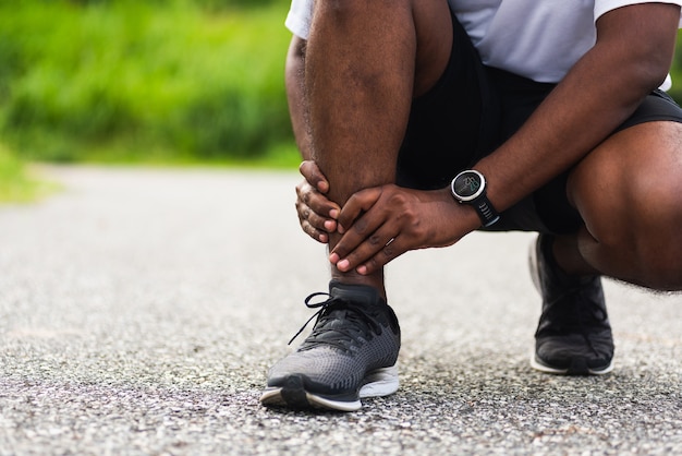 Молодой спортивный бегун черный мужчина носит часы, руки, суставы, боль в ноге из-за скрученной лодыжки, сломанной во время бега