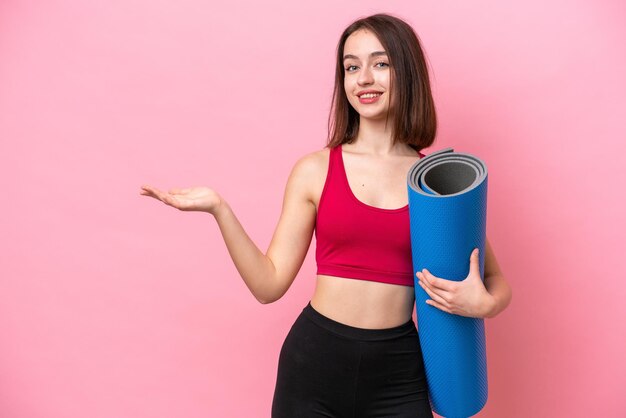 Young sport ukrainian woman going to yoga classes while holding\
a mat isolated on pink background extending hands to the side for\
inviting to come