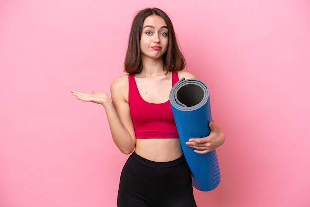 Young sport Ukrainian woman going to yoga classes while holding a mat isolated on pink background having doubts while raising hands