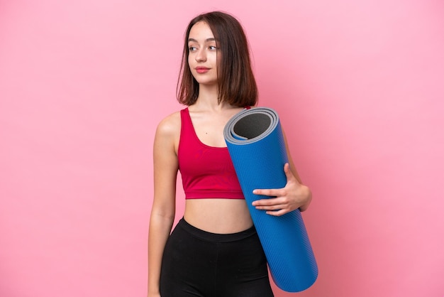 Young sport Ukrainian woman going to yoga classes while holding a mat isolated on pink background looking to the side