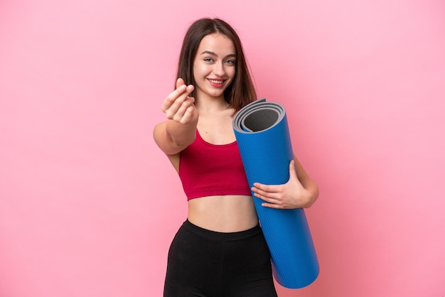 Young sport Ukrainian woman going to yoga classes while holding a mat isolated on pink background making money gesture