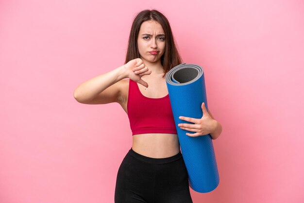 Young sport Ukrainian woman going to yoga classes while holding a mat isolated on pink background showing thumb down with negative expression