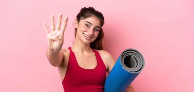 Фото Молодая спортивная женщина идет на занятия йогой, держа коврик счастливым и считая четыре пальцами