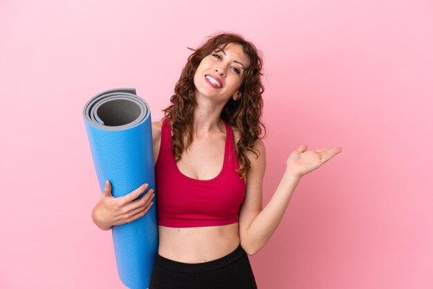 Young sport woman going to yoga classes while holding a mat isolated on pink background extending hands to the side for inviting to come