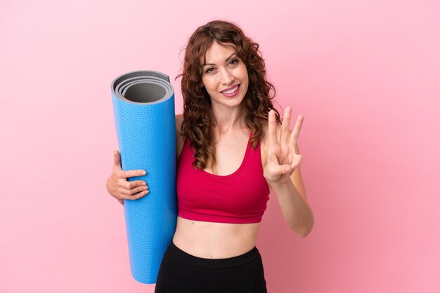 Young sport woman going to yoga classes while holding a mat isolated on pink background happy and counting three with fingers