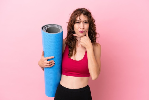 Young sport woman going to yoga classes while holding a mat isolated on pink background happy and smiling