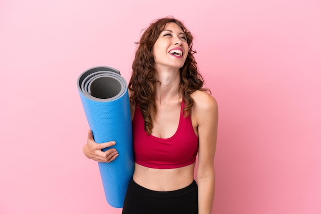 Young sport woman going to yoga classes while holding a mat isolated on pink background laughing