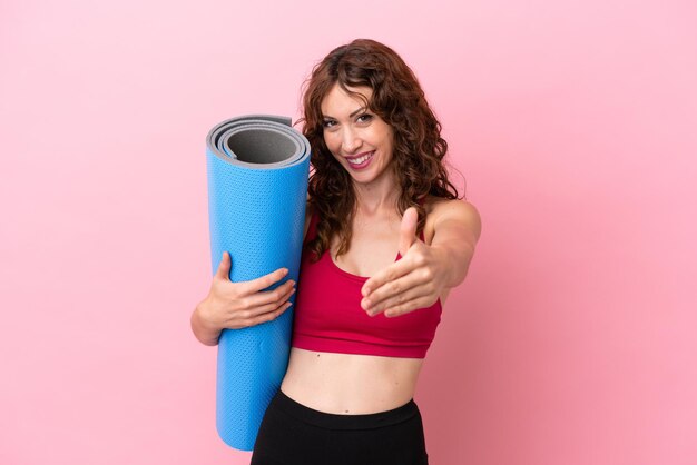 Young sport woman going to yoga classes while holding a mat isolated on pink background shaking hands for closing a good deal