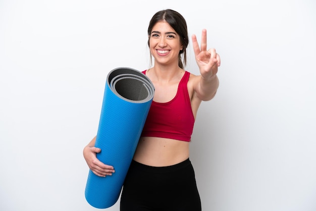 Young sport woman going to yoga classes while holding a mat isolated on white background smiling and showing victory sign