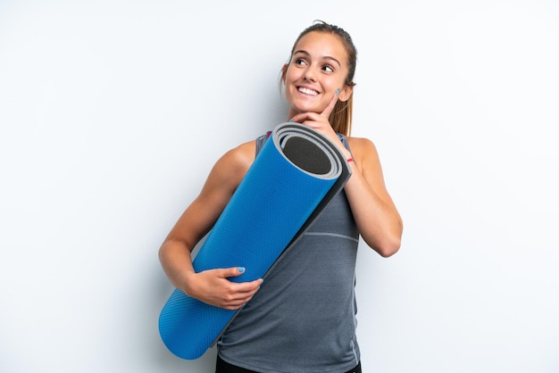 Giovane donna sportiva che va alle lezioni di yoga tenendo un tappetino isolato su sfondo bianco pensando un'idea mentre guarda in alto