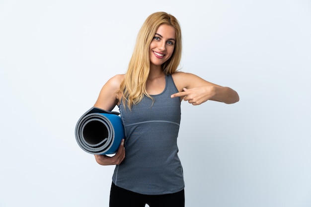 Young sport woman going to yoga classes while holding a mat over isolated white space and pointing it