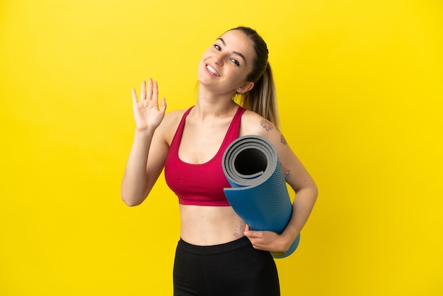 Young sport woman going to yoga classes while holding a mat saluting with hand with happy expression