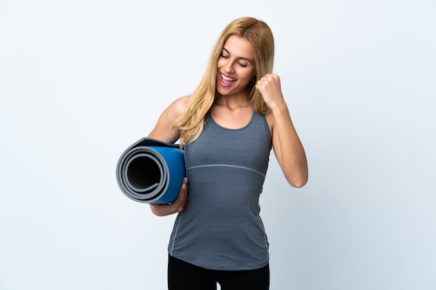 Young sport woman going to yoga classes while holding a mat over white wall celebrating a victory