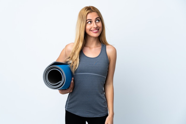 Young sport woman going to yoga classes while holding a mat over white wall looking up while smiling