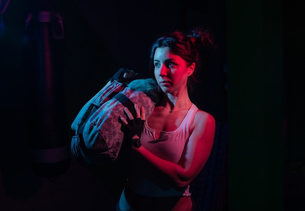 Young sport woman holding a heavy fitness bag for workout in neon gradient blue red light over a dark wall Functional Training Concept