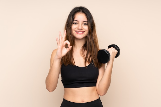 Young sport woman making weightlifting on beige wall showing an ok sign with fingers