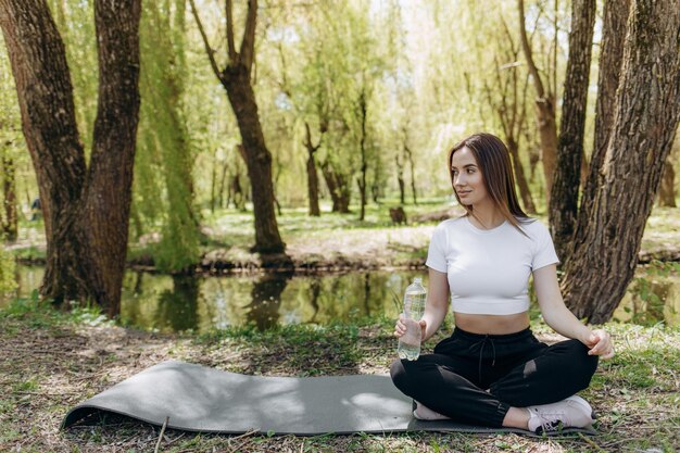 Young sport woman practice yoga on gymnastic mat in green park on grass Do morning breathing exercises in sportswear in summer nature background Concept of healthy lifestyle and harmony with body