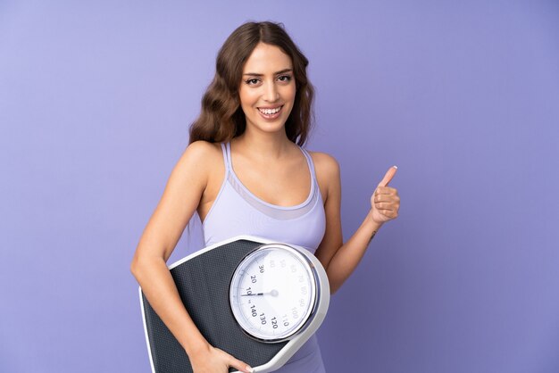 Young sport woman over purple wall holding weighing machine with thumb up