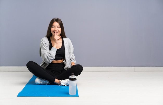 Young sport woman sitting on the floor with mat doing silence gesture