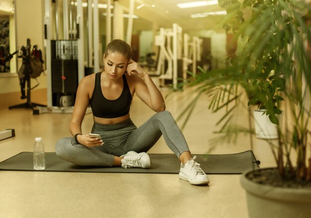 ジムでマットの上に座ってスマートフォンを使用してスポーツウェアの若いスポーツ女性。トレーニング休憩。健康的なライフスタイルのコンセプト