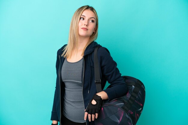 Young sport woman with sport bag isolated on blue background looking to the side