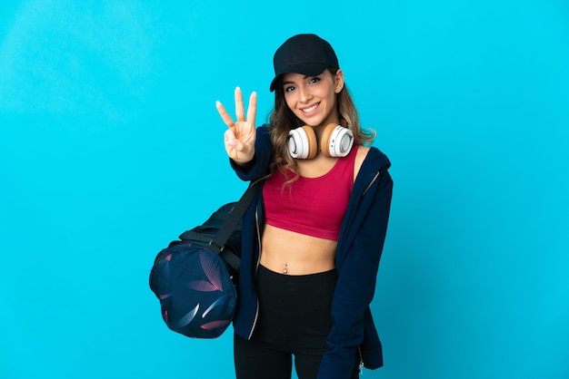 Young sport woman with sport bag isolated on blue wall happy and counting three with fingers