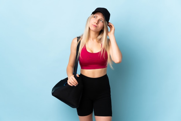 Young sport woman with sport bag isolated on blue wall having doubts