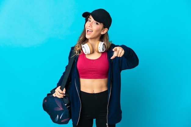 Young sport woman with sport bag isolated on blue wall surprised and pointing front